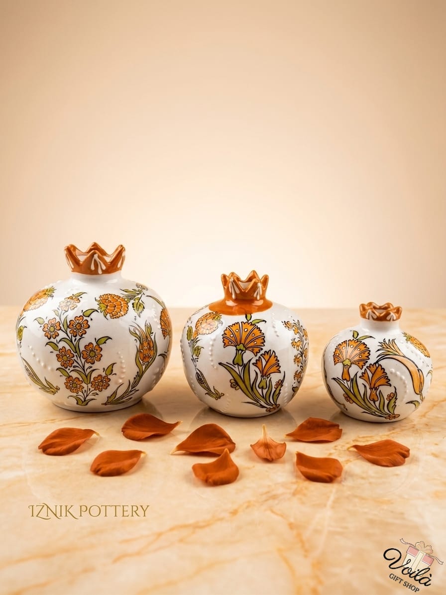 Set of three premium ceramic pomegranate vases in cascading sizes, featuring elegant orange and green traditional Iznik floral patterns with a glazed amber scalloped crown, surrounded by scattered orange petals.
