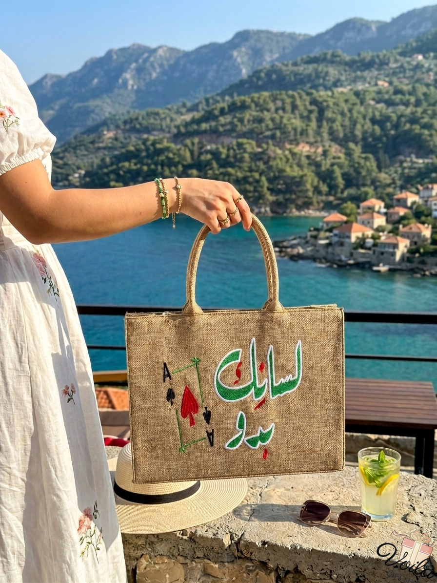 Handcrafted jute tote bag with Lebanese slang embroidery held against a coastal Mediterranean background