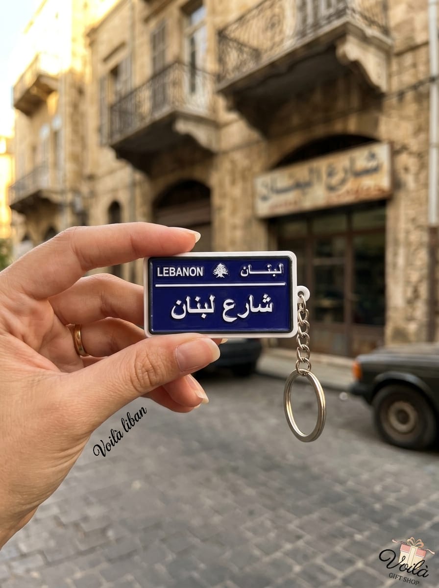 Hand holding a miniature blue Lebanese street sign keychain reading Lebanon Street in Arabic (Share' Loubnan), set against a traditional cobblestone street and classic architecture.