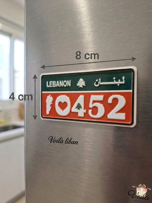 Red and green decorative Lebanese license plate fridge magnet measuring 8cm by 4cm, displaying the patriotic number 10452 with a heart, the national Cedar tree, and the white silhouette of the Lebanon map, placed on a stainless steel refrigerator.