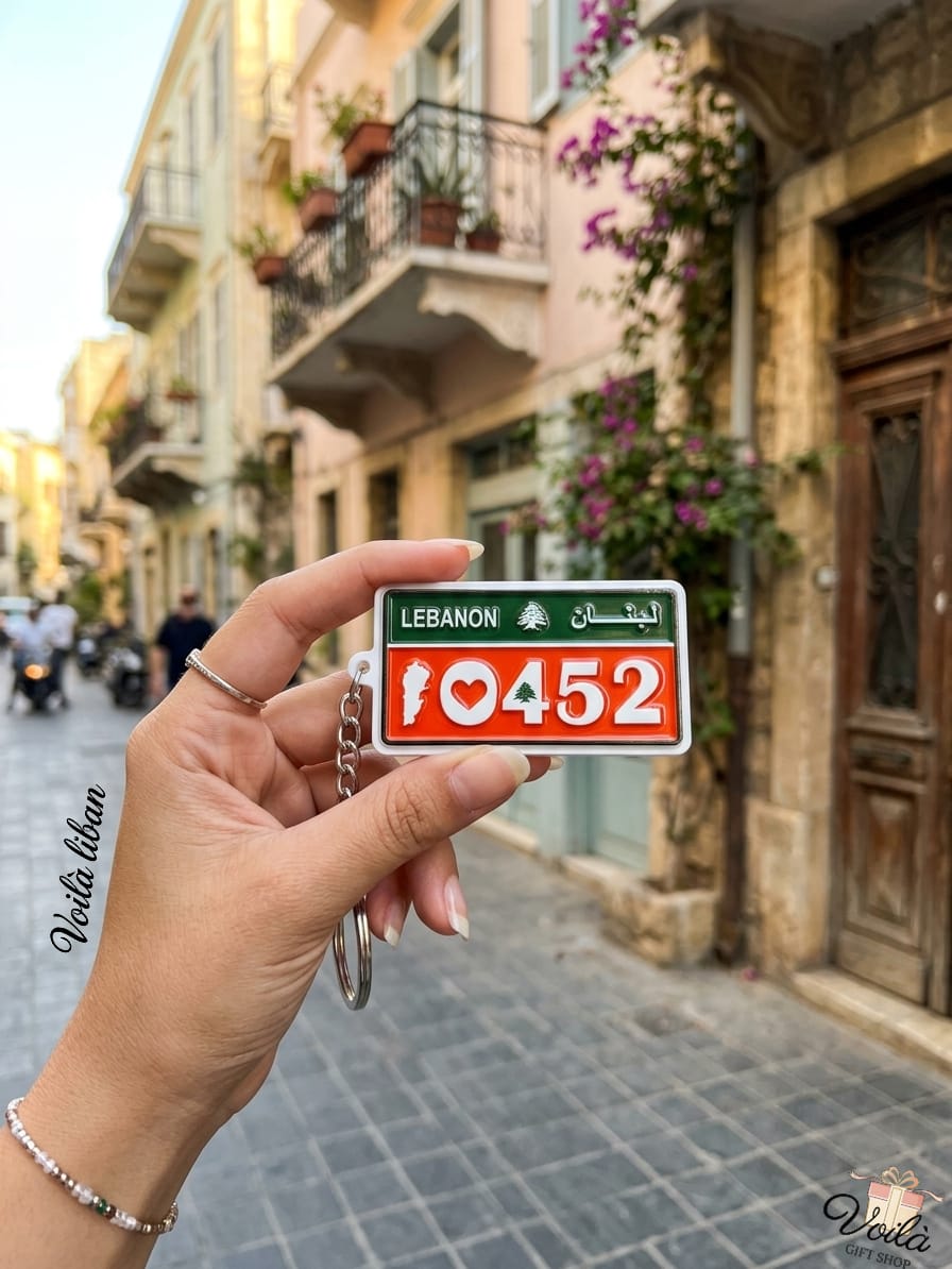 Hand holding a miniature red and green Lebanese license plate keychain displaying the patriotic number 10452 with a heart, the national Cedar tree, and the Lebanon map silhouette, against a traditional street backdrop.