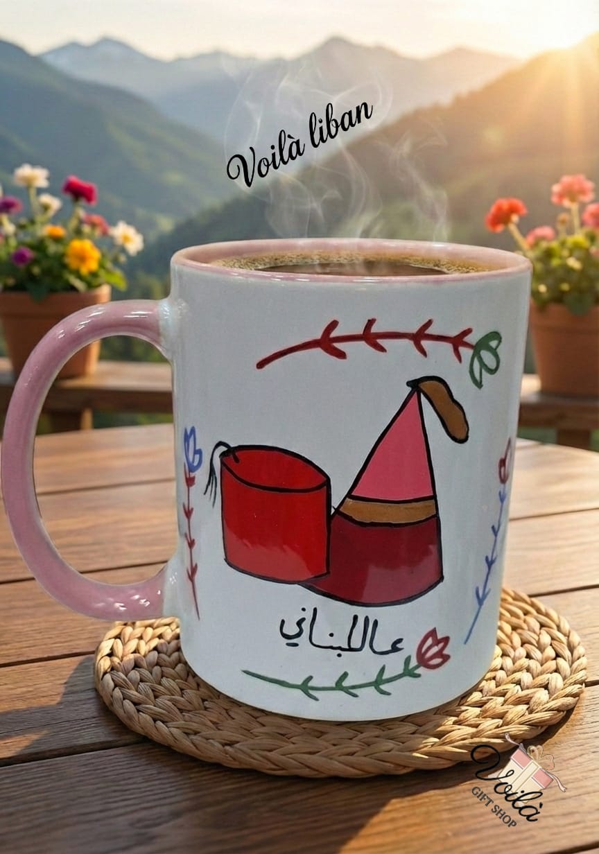 White mug with pink handle on a wooden table, featuring a design and text, with a scenic background.