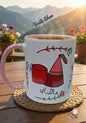 White mug with pink handle on a wooden table, featuring a design and text, with a scenic background.