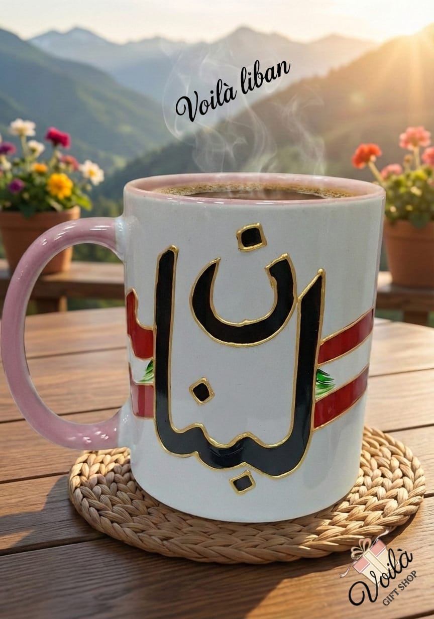 White mug with Arabic text and pink handle on a wooden table with a mountainous background