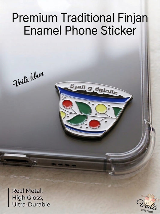 Premium metal enamel phone sticker shaped like a traditional Lebanese coffee finjan with a classic floral pattern in green, red, and yellow, featuring the Arabic phrase "عالحلوة والمرة" written along the top rim, attached to a clear phone case.