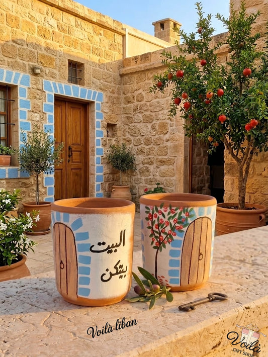 Two hand-painted terracotta mugs sitting on a stone ledge next to a vintage key. Both mugs feature a traditional wooden door with blue stone trim against a white background. One mug has the Arabic phrase Al Bayt Baytkun, while the other features a painted pomegranate tree. A traditional Lebanese courtyard is in the background.