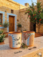 Two hand-painted terracotta mugs sitting on a stone ledge next to a vintage key. Both mugs feature a traditional wooden door with blue stone trim against a white background. One mug has the Arabic phrase Al Bayt Baytkun, while the other features a painted pomegranate tree. A traditional Lebanese courtyard is in the background.