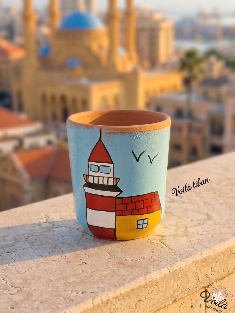 Hand-painted terracotta mug featuring the iconic red and white Al Manara lighthouse with a small yellow building against a light blue sky with flying birds, sitting on a stone balcony overlooking a blurred Beirut skyline.