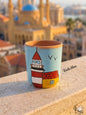 Hand-painted terracotta mug featuring the iconic red and white Al Manara lighthouse with a small yellow building against a light blue sky with flying birds, sitting on a stone balcony overlooking a blurred Beirut skyline.
