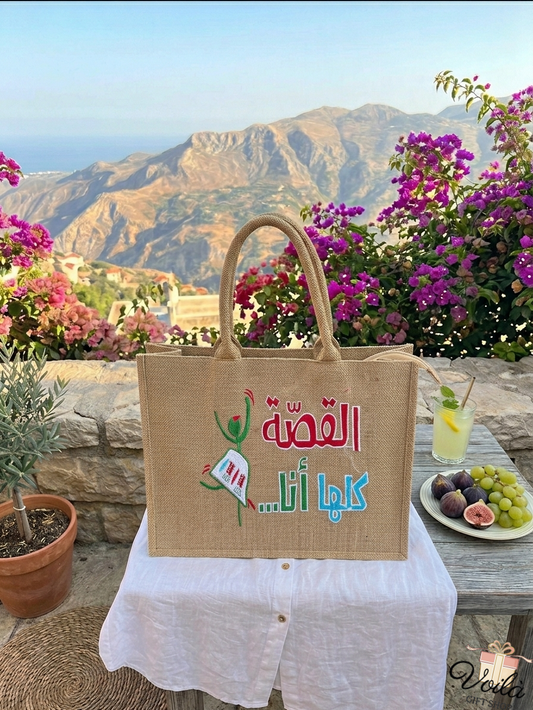 Al Qissa Kella Ana jute tote bag with Arabic calligraphy and embroidered design, set against a beautiful Lebanese mountain and sea landscape with bougainvillea flowers.