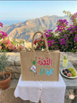 Al Qissa Kella Ana jute tote bag with Arabic calligraphy and embroidered design, set against a beautiful Lebanese mountain and sea landscape with bougainvillea flowers.