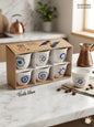 Set of six authentic white ceramic Arabic coffee cups featuring assorted blue protective evil eye motifs, presented in a premium kraft display box on a white marble countertop next to a copper rakwe.