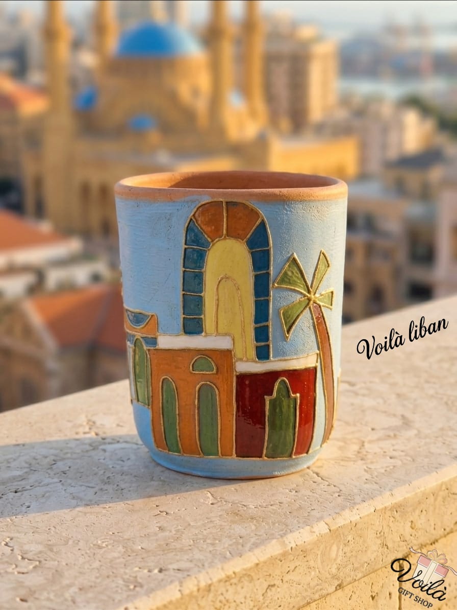 Hand-painted terracotta mug featuring a stylized, vibrant Beirut cityscape with traditional colorful arches, buildings, and a palm tree, sitting on a stone balcony with a blurred view of the Mohammad Al-Amin Mosque in the background.