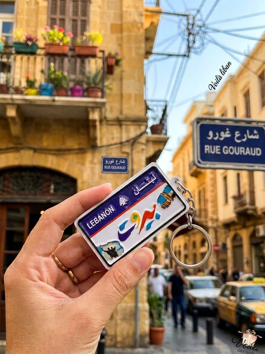 Hand holding a miniature Beirut Lebanon license plate keychain featuring Raouche Rocks and Martyrs' Monument, with a classic Beirut street and Rue Gouraud sign in the background.