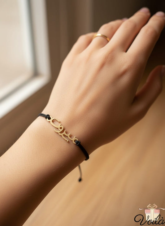 Hand wearing a gold bracelet with a black cord on a wooden surface