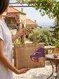 Hand-painted jute tote bag featuring a chic woman in a purple hat, held in front of a traditional Lebanese stone house