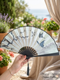 Hand holding an elegant silk folding fan with butterfly and blossom design on a sunny terrace