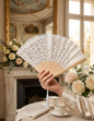 Hand holding an elegant white lace folding fan with a wooden frame and white tassel