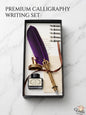 Premium calligraphy writing set with purple feather, gold pen, ink bottle, and paper in a black box on a marble background.