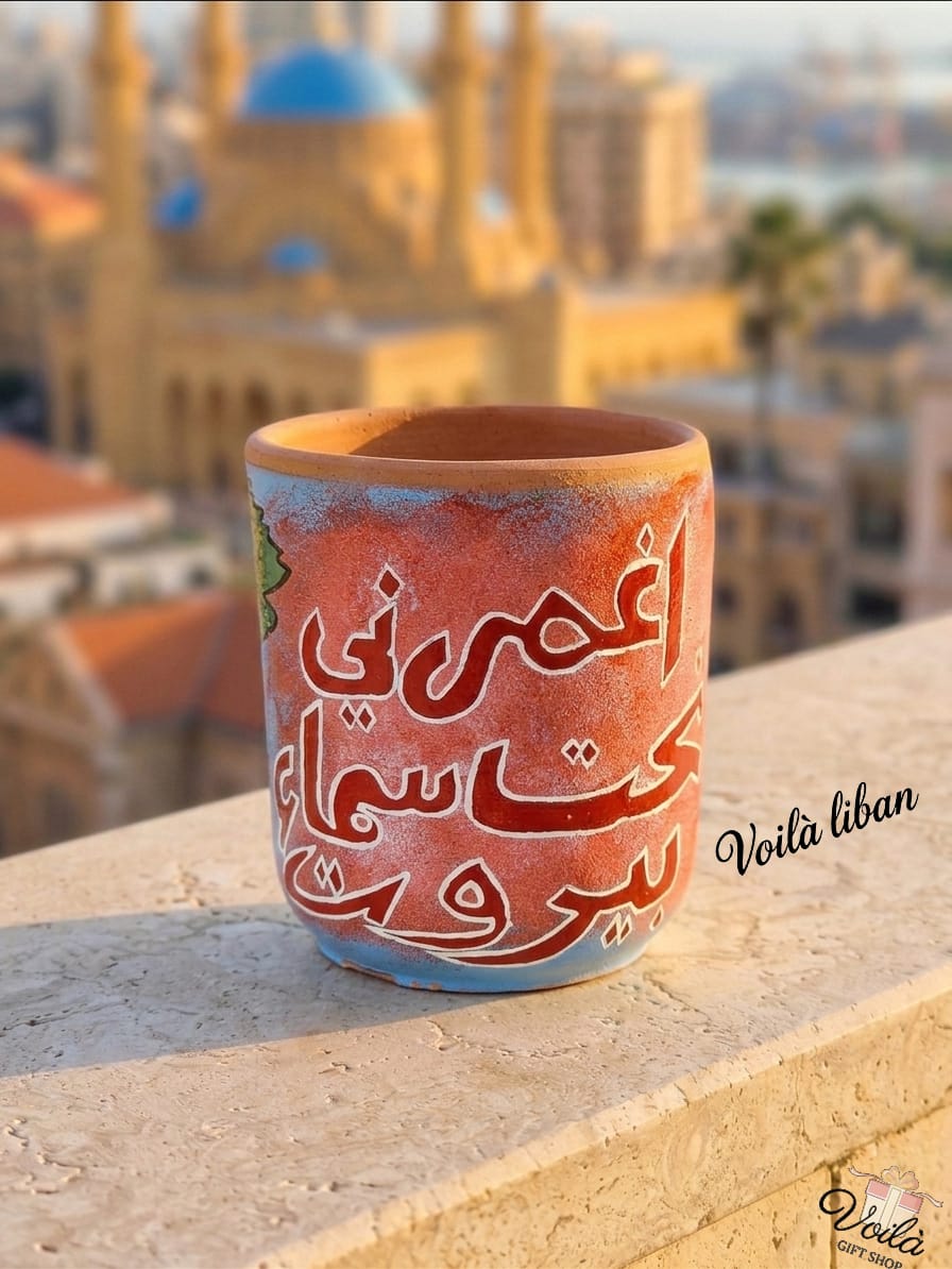 Hand-painted terracotta mug in sunset orange and light blue, featuring the Arabic calligraphy phrase Ghmorni Tahet Sama Beirut, resting on a stone balcony with a blurred Beirut skyline and blue-domed mosque in the background.
