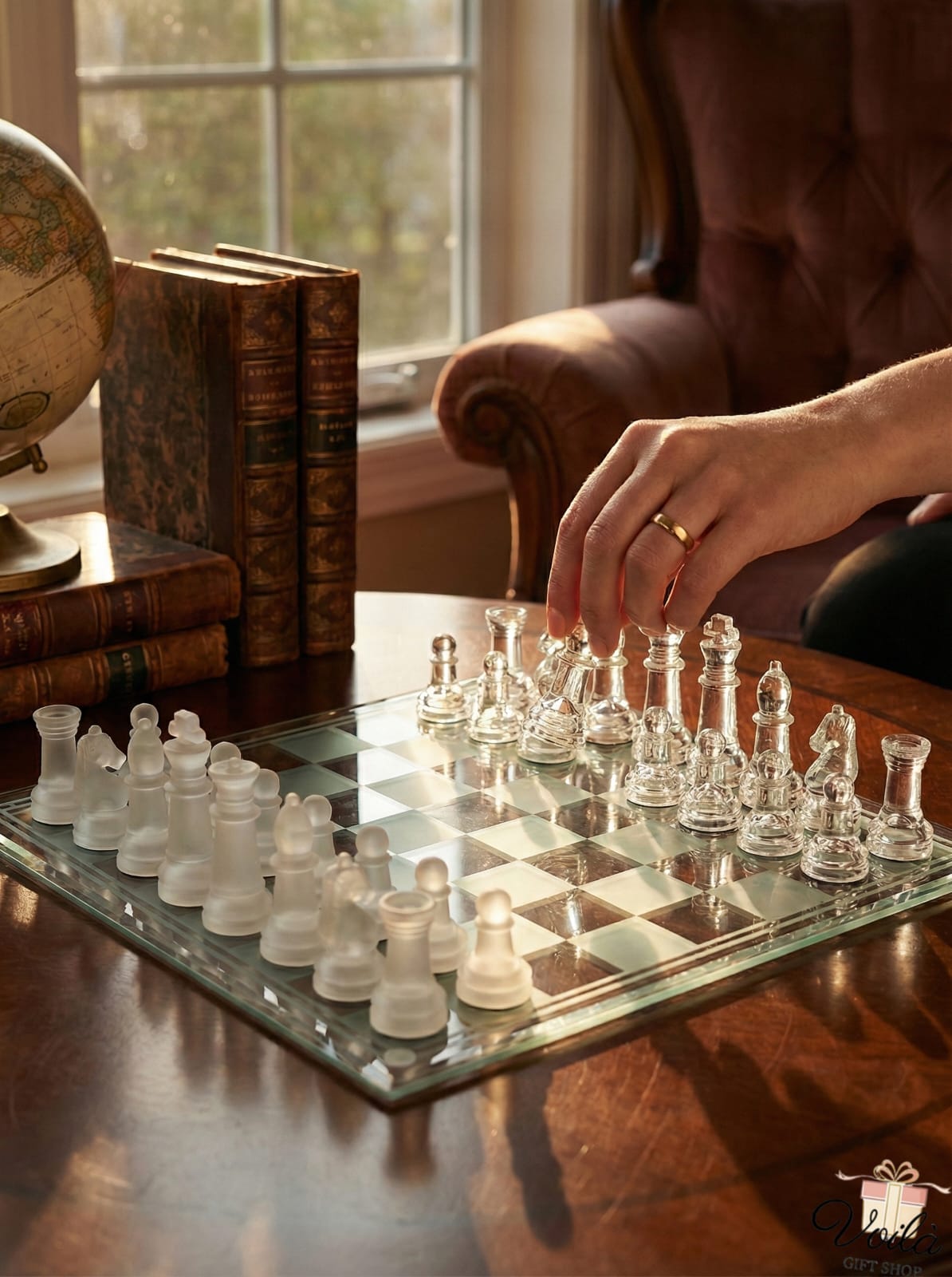 Clear glass chess pieces on a reflective chessboard with a hand making a move, in a room with books and a globe.