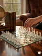 Clear glass chess pieces on a reflective chessboard with a hand making a move, in a room with books and a globe.