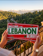 Vintage Greetings from Lebanon metal license plate sign held against a scenic mountain backdrop