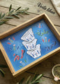 Decorative tray with a blue background featuring white cups and green leaves, placed on a wooden surface.