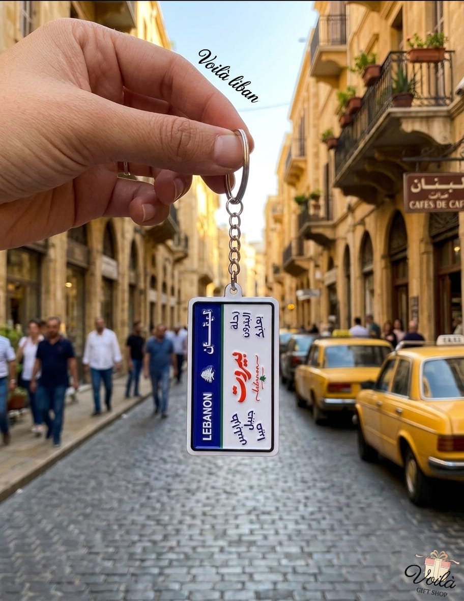 Hand holding a miniature vertical Lebanese license plate keychain featuring the names of iconic cities in Arabic calligraphy including Beirut in bold red, with a lively Lebanese street scene and yellow taxi in the background.