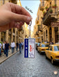 Hand holding a miniature vertical Lebanese license plate keychain featuring the names of iconic cities in Arabic calligraphy including Beirut in bold red, with a lively Lebanese street scene and yellow taxi in the background.