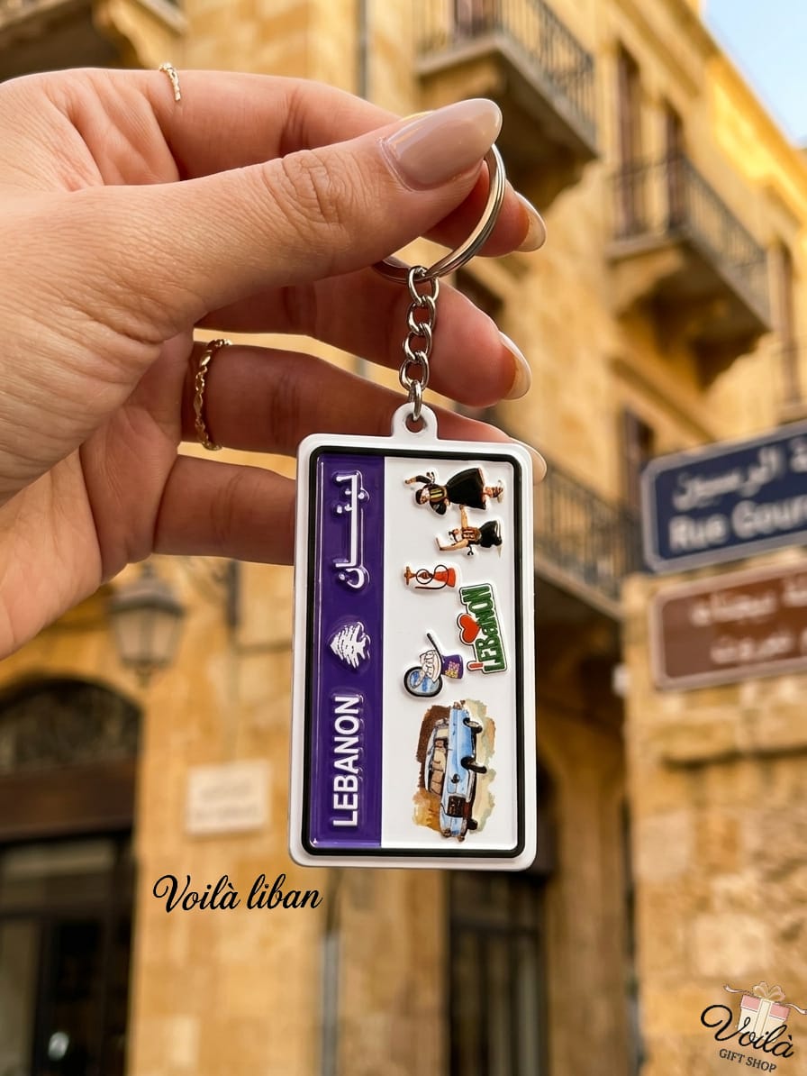 Hand holding a miniature vertical Lebanese license plate keychain featuring cultural icons including a vintage blue service taxi, traditional Dabke dancers, a coffee rakwe, and a hookah, with a classic stone street in the background.
