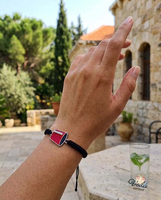 Handcrafted Lebanese heritage cord bracelet worn on a wrist with a traditional Lebanese stone house in the background.