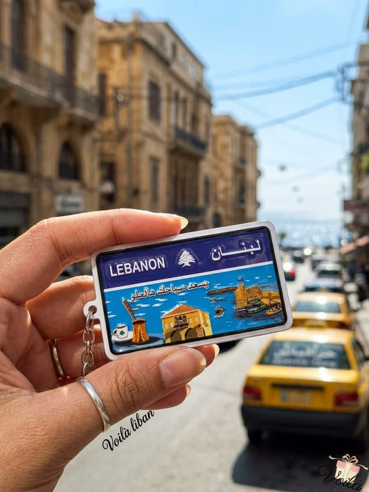 Hand holding a miniature blue Lebanese license plate keychain featuring a coastal scene with the Saida Sea Castle, traditional coffee, and the Arabic phrase Yis'ed sabahak ya helo, with classic yellow taxis in a sunny street background.