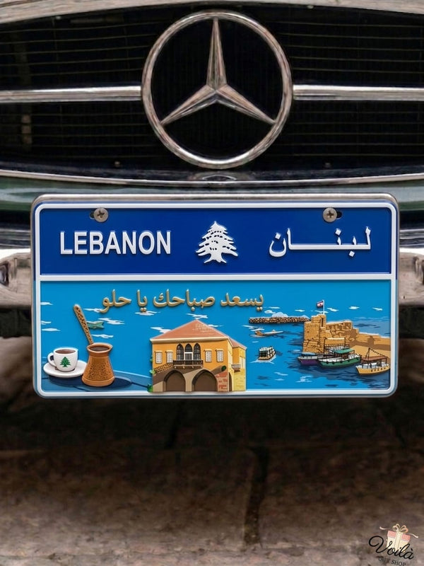 Decorative blue Lebanese license plate mounted on a classic car, featuring a bright coastal scene with the Saida Sea Castle, a traditional stone house, a coffee rakwe, and the Arabic phrase Yis'ed sabahak ya helo.