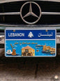 Decorative blue Lebanese license plate mounted on a classic car, featuring a bright coastal scene with the Saida Sea Castle, a traditional stone house, a coffee rakwe, and the Arabic phrase Yis'ed sabahak ya helo.