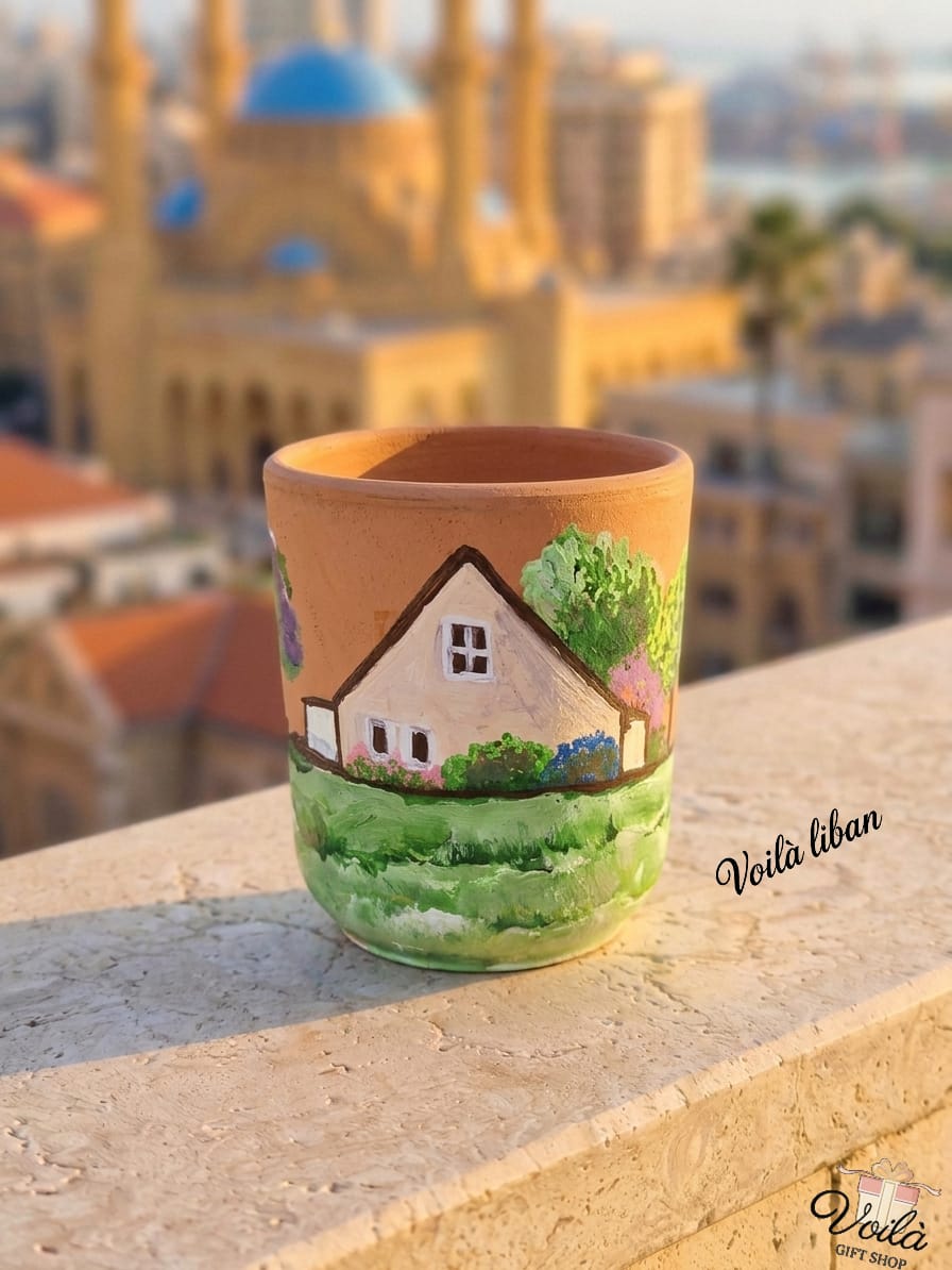 Hand-painted terracotta mug featuring a traditional Lebanese mountain house with a pitched roof, surrounded by lush green grass and blooming trees, resting on a stone balcony with a blurred Beirut skyline in the background.