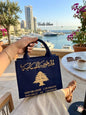 Woman holding a premium navy blue tote bag with gold embroidered Lebanese Republic cedar tree design, overlooking a scenic marina in Beirut.