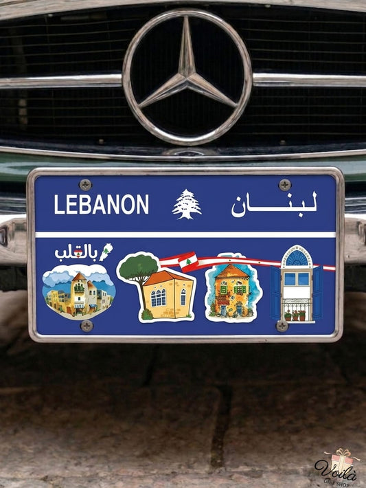 Blue decorative Lebanese license plate mounted on a classic car, featuring illustrations of traditional village houses with red tile roofs, iconic triple-arched windows, the Lebanese flag, and Arabic text reading Bil Qaleb.