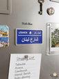 Miniature blue Lebanese street sign fridge magnet reading Lebanon Street in Arabic (Share' Loubnan), placed on a stainless steel refrigerator holding a Lebanese food shopping list.