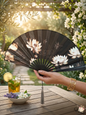 Elegant black silk hand fan with white lotus pattern held in a sunny garden setting
