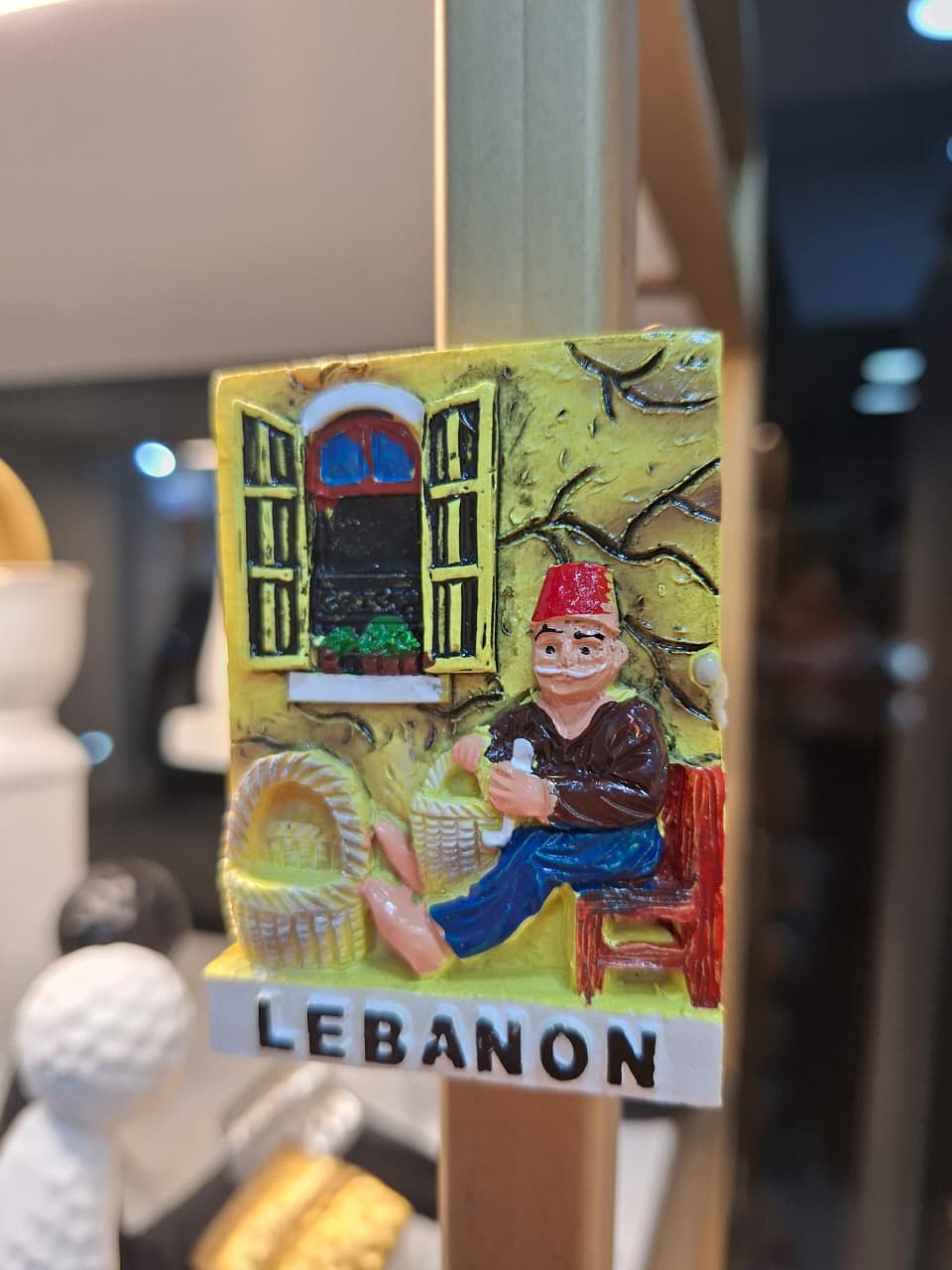 Decorative magnet with a man sitting on a chair, holding a basket, and a window in the background, labeled 'Lebanon'.