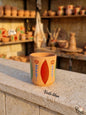 Hand-painted terracotta mug featuring a traditional folk art floral design with a red geometric center and blue stems with red tulips, sitting on a stone ledge with a blurred pottery workshop in the background.