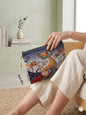 Woman sitting in a bright room holding a premium woven tapestry pouch featuring a vibrant, patterned folk-art style cat among wildflowers against a dark starry night background.