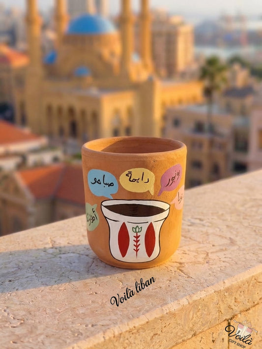 Hand-painted terracotta mug featuring a traditional Lebanese coffee finjan surrounded by colorful speech bubbles with Arabic morning greetings like Sabaho, Ray2a, and Bonjour, sitting on a stone balcony with a blurred Beirut skyline.