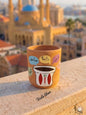 Hand-painted terracotta mug featuring a traditional Lebanese coffee finjan surrounded by colorful speech bubbles with Arabic morning greetings like Sabaho, Ray2a, and Bonjour, sitting on a stone balcony with a blurred Beirut skyline.