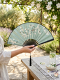 Elegant green silk folding hand fan with white blossoms and black tassel held in a sunny garden setting.