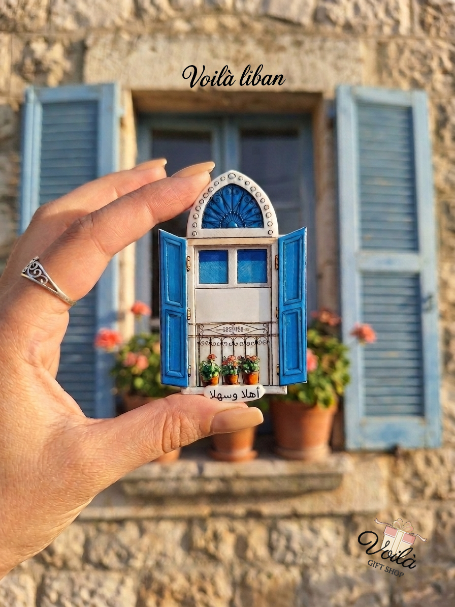 Hand holding a miniature traditional Lebanese blue window magnet with Ahla w Sahla text in front of a real stone house window