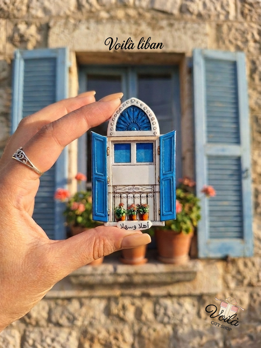 Hand holding a miniature traditional Lebanese blue window magnet with Ahla w Sahla text in front of a real stone house window