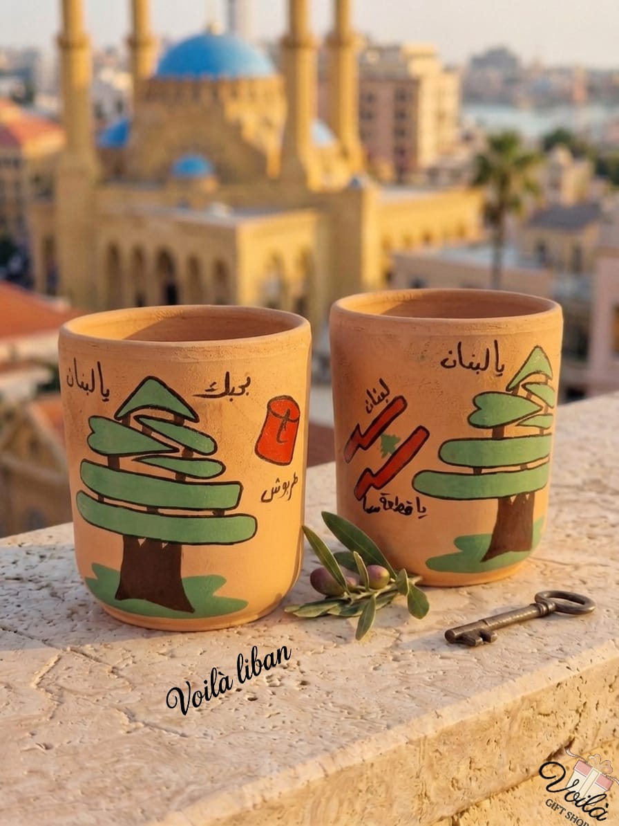 Two hand-painted terracotta mugs on a stone ledge overlooking a blurred Beirut skyline. Both feature a green Lebanese cedar tree; one includes a traditional red tarboush with the text Ya Libnan Bhebak, and the other includes a stylized red Lebanon flag with the text Ya Libnan Ya Eteet Sama.